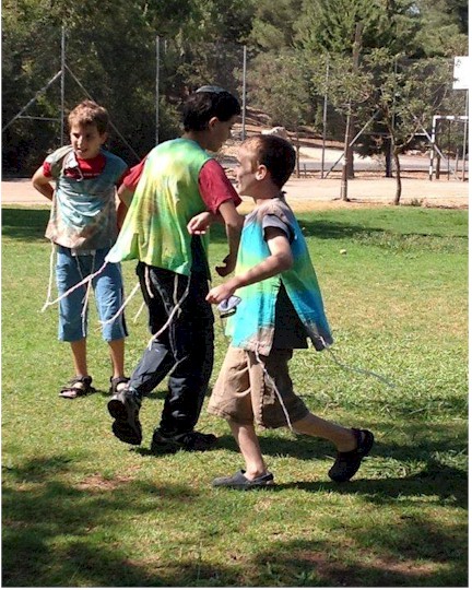 Three smiling children proudly wearing colorful tie-dye tzitzit